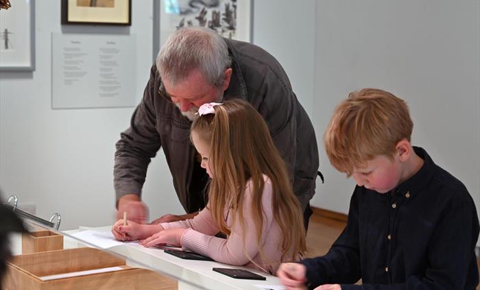 Man and 2 children (boy and girl) doing the rubbings activity at 'The Lost Words' exhibition